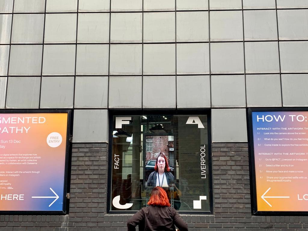 Lesley standing outside FACT Liverpool, her face reflected in the glass entrance between colourful Augmented Empathy exhibition signage