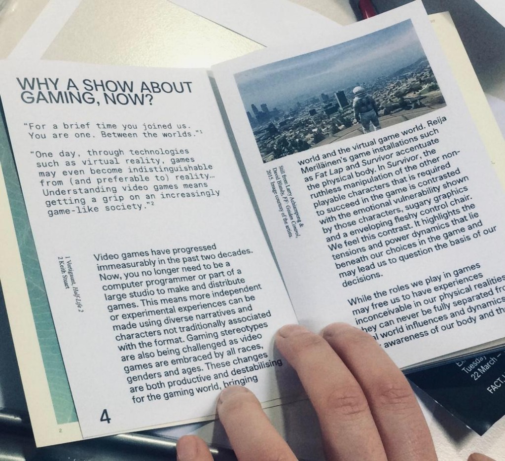 Hand holding open a printed exhibition zine titled 'Why A Show About Gaming, Now?' with essay text and a video game screenshot