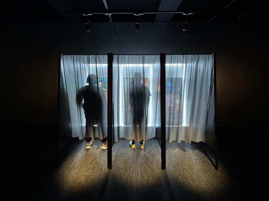 Two figures silhouetted behind translucent curtains in a darkened gallery space, backlit by strip lights across three curtained booths