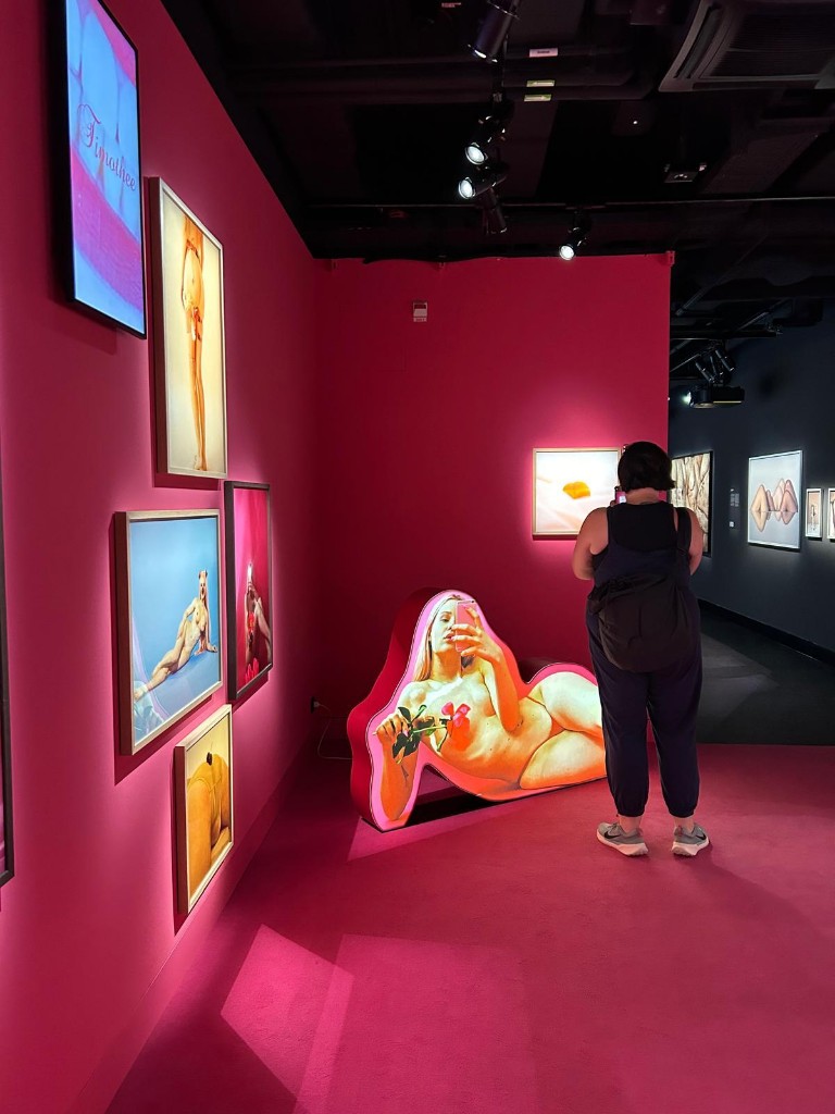 Visitor viewing artwork in a vivid pink gallery room with framed photographs on the wall and a large body-shaped cutout sculpture on the floor